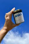 a hand coming out of the side of the frame, lit from the sun to the left, holding a hawaiian macadamia nut butter jar in a blue spirulina flavor with a gold lid and the maka logo and packaging design, against a background of blue sky with clouds