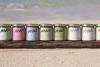 maka hawaiian macadamia nut butter jars in various flavors, photographed side by side on a wooden board resting on a sandy beach with the tide in the background