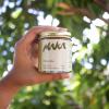 a hand coming out the side of the frame holding a hawaiian macadamia nut butter jar with a gold top the maka logo and packaging design in front of an unfocused background of foliage