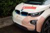 Front view of a BMW i3 in front of a green hedge wrapped in graphics for Pinecone Childcare in Portland, Oregon. It is mostly beige with minty green and pink shapes. It has the pinecone logo on it and a red shape with a qr code and text that reads "Register now!" and "pinecone-childcare.com".