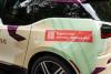 Back left view of a BMW i3 wrapped in graphics for Pinecone Childcare in Portland, Oregon. It is beige with minty green and dark purple shapes. It features a red shape with a qr code and text that reads "Register now!" and "pinecone-childcare.com".